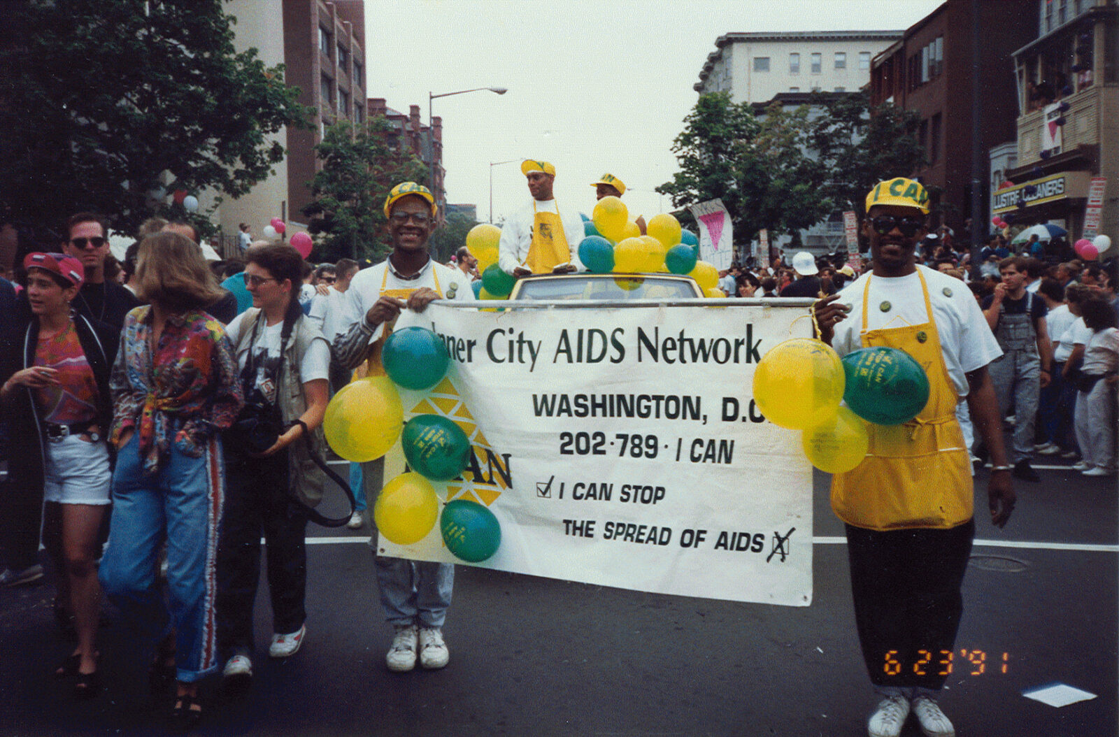 A photograph of two people smiling and holding up a banner that reads “Inner City AIDS Network Washington, D.C.” with bunches of yellow and blue balloons. Behind them are throngs of marchers and a car with more balloons on top.