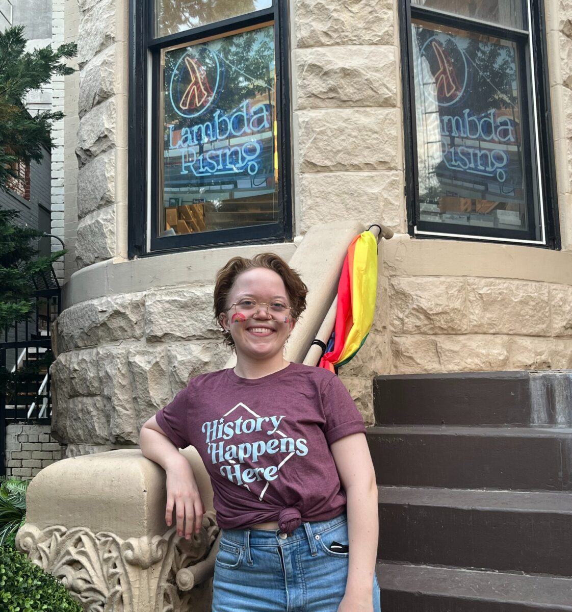 Image description: Emma, a white person with short brown hair and glasses, stands on the steps in front of a building with a photo of the “Lambda Rising” neon sign in its window. Emma is wearing a t-shirt that reads “History Happens Here” with an outline of the shape of DC.