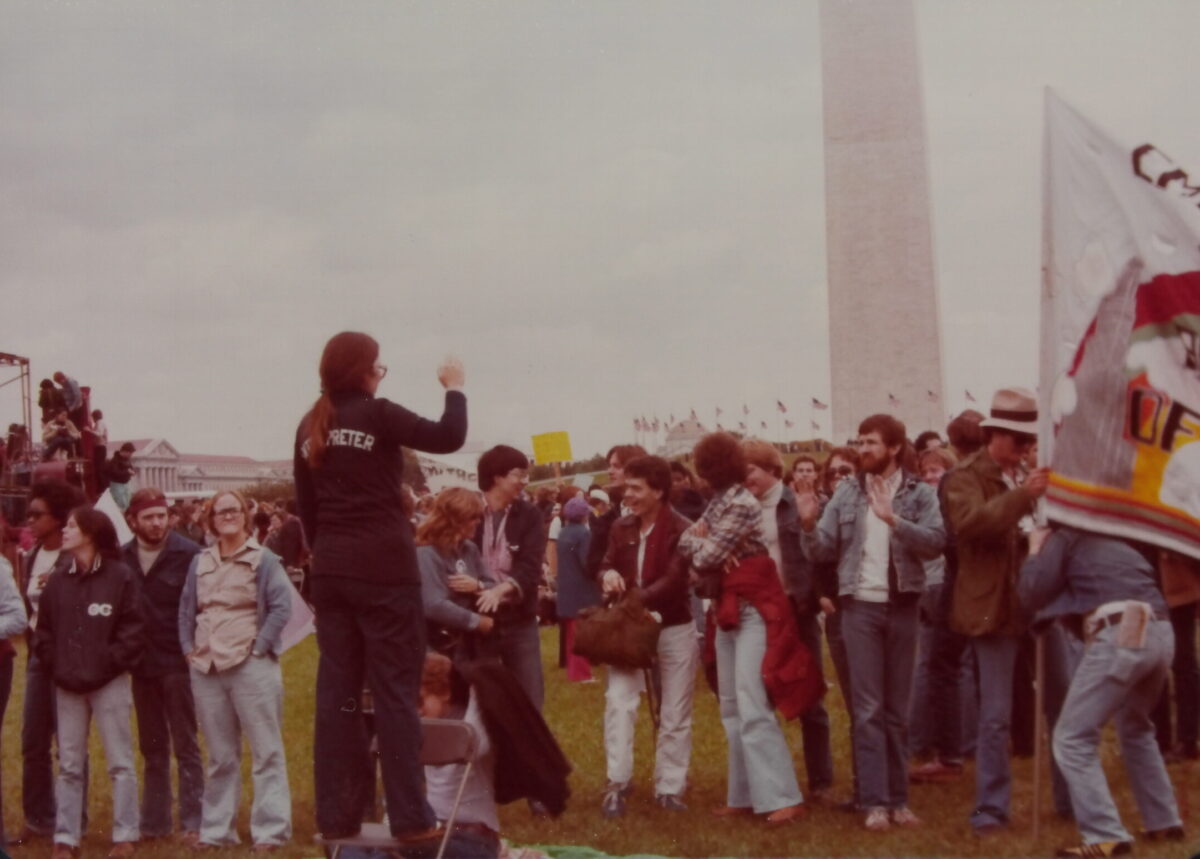 A photo of a group of people gathered at the foot of the Washington monument. They are all standing around a person standing on a chair wearing a jacket that says "interpreter" across the back. She is signing to the crowd.